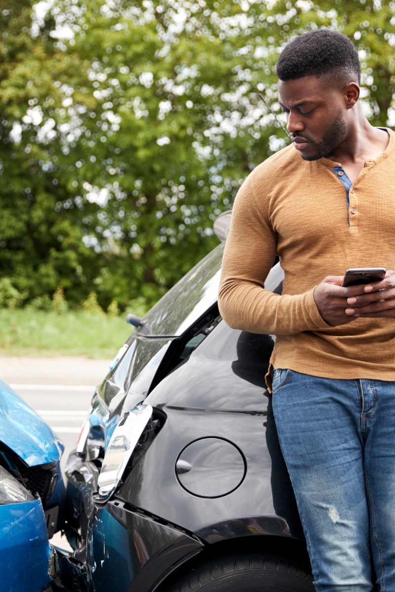 A man inspecting his phone near a damaged car.
