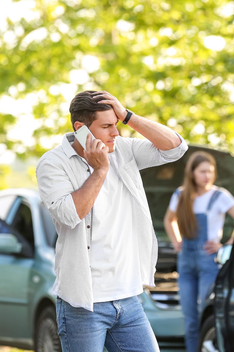 A man standing next to a broken down car while talking on a cell phone.