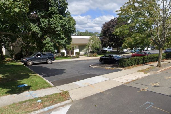 A small parking lot with several cars parked, surrounded by trees and a low white building in the background on a partly cloudy day in East Islip NY, near trusted Workers Compensation doctors.