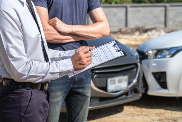 A man standing next to a car with a clipboard in front of it, possibly working as an insurance adjuster or accident investigator for no-fault claims.