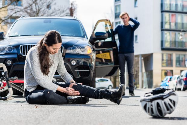 A woman, injured in a car accident, sits on the ground next to a broken-down car.