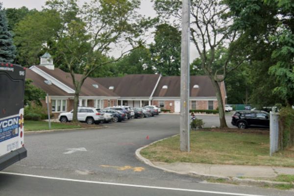 A parking lot filled with several cars lies before a single-story brick building, perhaps a doctor’s office, surrounded by trees. On the left, a bus is partially visible.