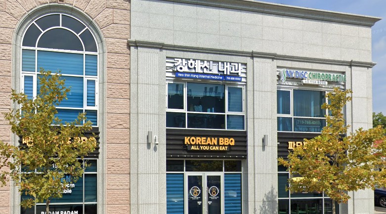 Two-story commercial building in Flushing NY with signs for a Korean BBQ restaurant, a chiropractic clinic, and a business with Korean text. Three small trees are visible in front—home to Workers Compensation Doctors and more.