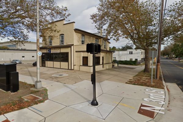 A beige two-story building with a business sign stands on the corner of N 32nd St., trees lining the sidewalk, and a streetlight in the foreground. This serene Cherry Hill NJ location is ideal for No-Fault Doctors catering to workers' compensation needs in a calm and inviting atmosphere.