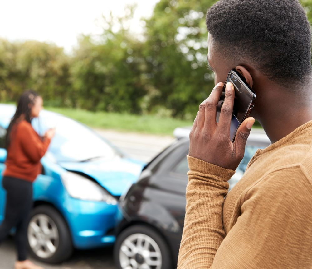 A man on the phone stands near a car accident involving two vehicles, possibly seeking advice from injury doctors in New Jersey. A woman in the background is also using her phone.