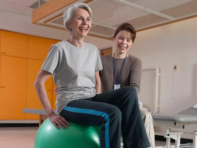 Older woman sitting on a green exercise ball with assistance from a younger person in a rehabilitation setting.