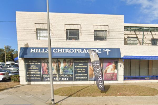 Street view of Hills Chiropractic, a two-story cream-colored building with a blue awning in Hempstead, NY. There are large window posters advertising various treatments, including Workers Compensation Doctors, and a flag reading "4200 Services" outside.