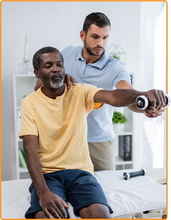 In New York, a therapist under the Workers' Comp program assists a man in an exercise, helping him lift a dumbbell. The man sits on a table, wearing a yellow shirt and dark shorts. In New York, a therapist under the Workers' Comp program assists a man in an exercise, helping him lift a dumbbell The man sits on a table, wearing a yellow shirt and dark shorts